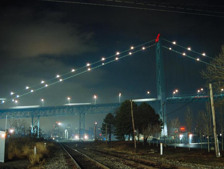 A suspension bridge with lights is visible over railroad tracks and trees at night.
