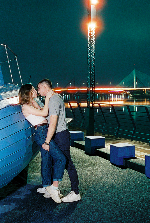 A couple embraces next to a blue structure on a riverside promenade at night, with a bridge and city lights in the background.