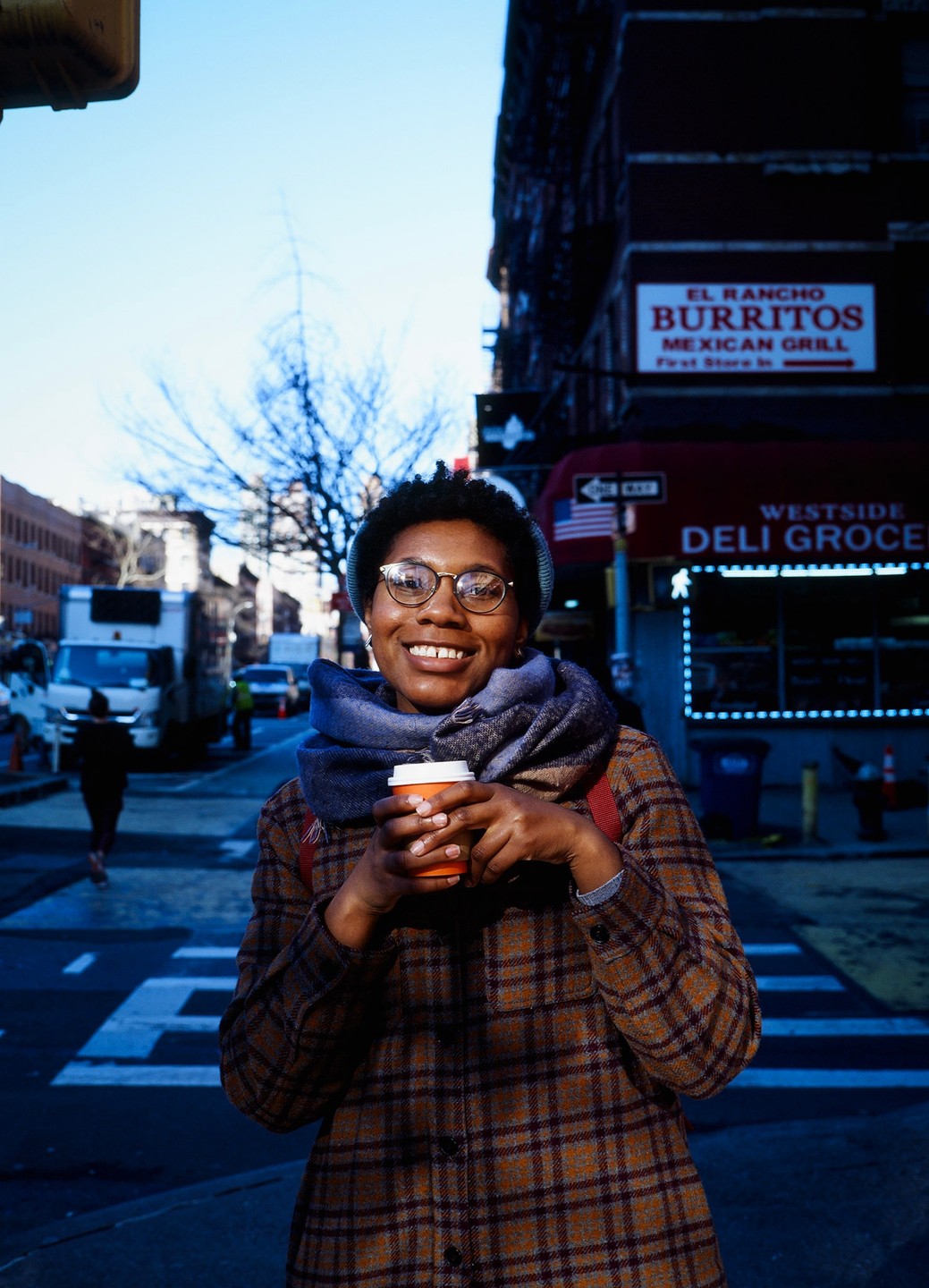A woman wearing a plaid coat and scarf smiles while holding a coffee cup on a city street.