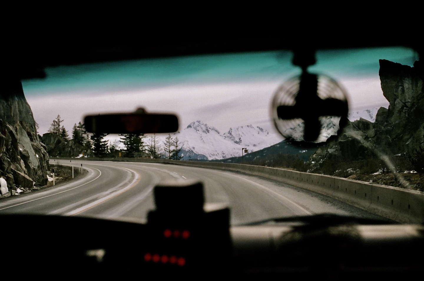 A winding road leads through a mountainous landscape with snow-capped peaks visible in the distance, viewed from inside a vehicle.