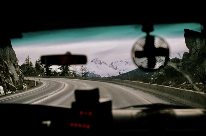 A winding road leads through a mountainous landscape with snow-capped peaks visible in the distance, viewed from inside a vehicle.