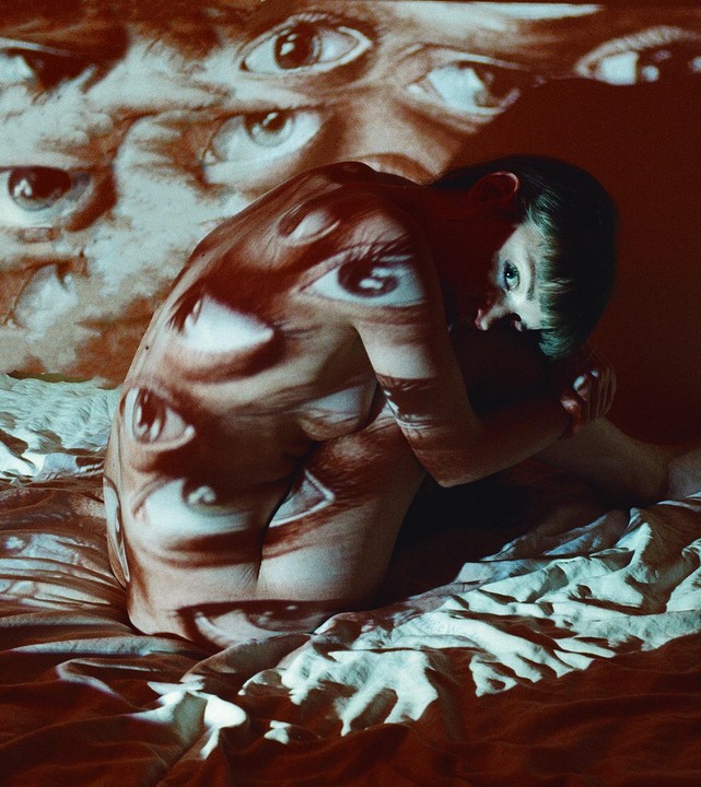 A woman sits on a bed with multiple eyes projected onto her body and the wall behind her.