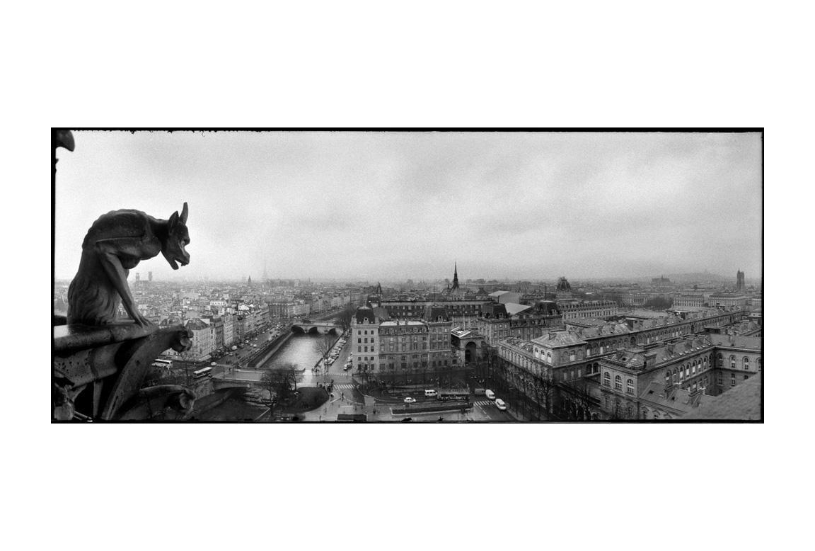 A gargoyle statue overlooks a panoramic view of a city with a river and many buildings under a cloudy sky.