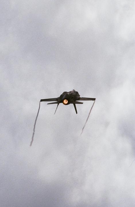 A jet aircraft with an illuminated engine exhaust flies through a cloudy sky, leaving two trails of smoke.