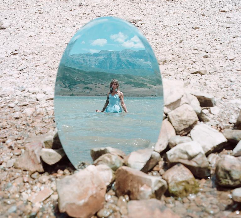 A woman stands in water, reflected in an oval mirror on a rocky shore with mountains in the background.