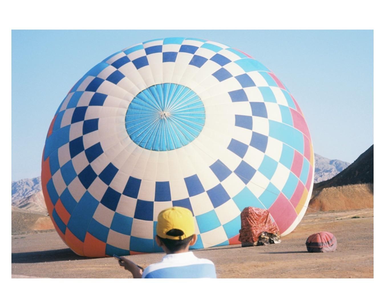 A person in a yellow hat stands in front of a large, deflated hot air balloon with a checkered pattern.