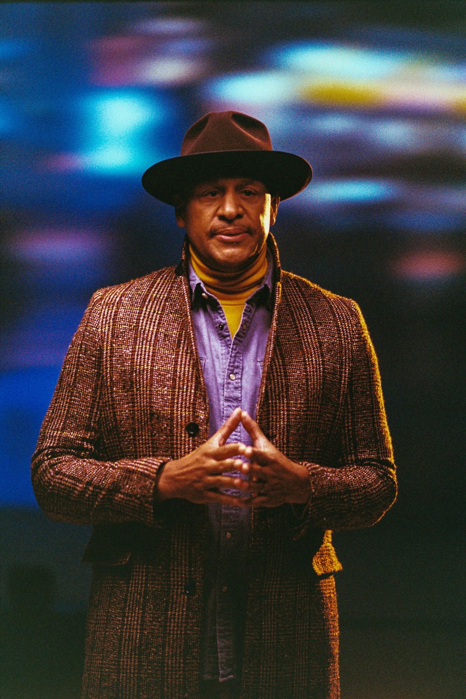 A man in a brown hat, yellow turtleneck, and plaid coat stands with his hands clasped in front of a blue and purple background.