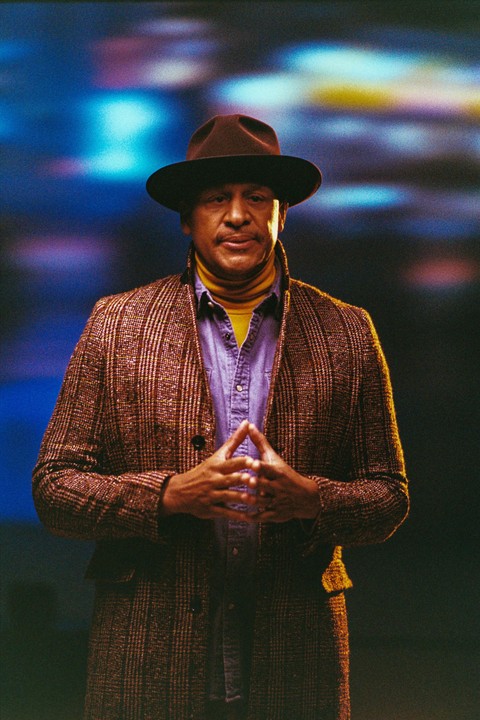 A man in a brown hat, yellow turtleneck, and plaid coat stands with his hands clasped in front of a blue and purple background.