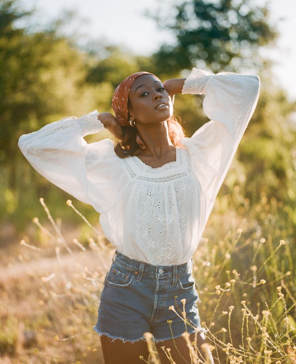 A woman wearing a white blouse, denim shorts, and a headscarf poses in a field of tall grass and small yellow flowers.