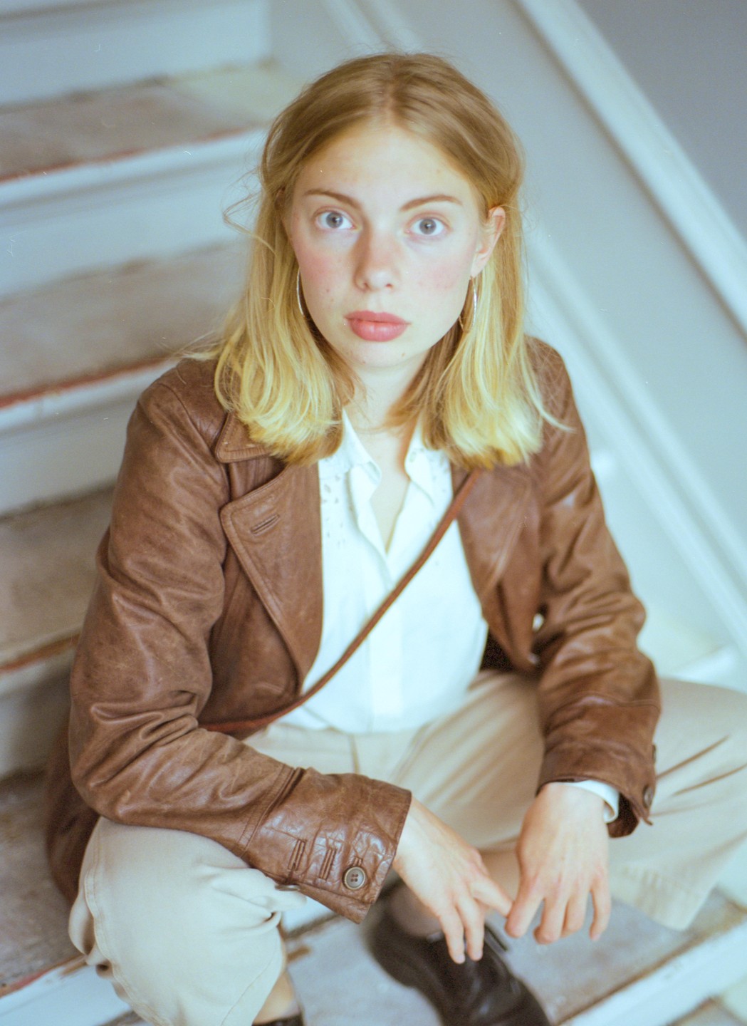 A woman with blonde hair and a brown leather jacket sits on a staircase.