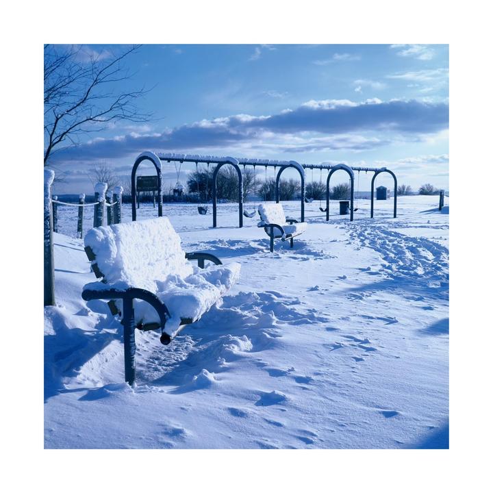 A snow-covered park bench sits in the foreground of a snowy playground with swings under a blue sky.