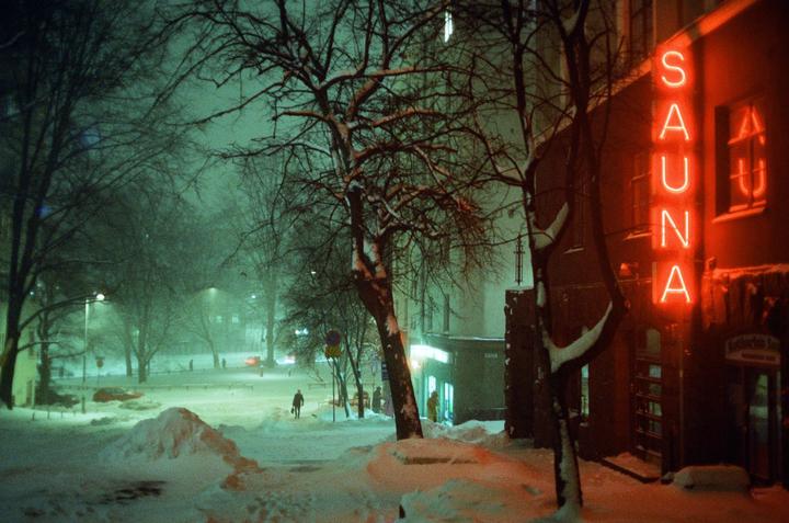 A neon sign for a sauna glows red on a snowy street at night, with bare trees and a person walking in the distance.