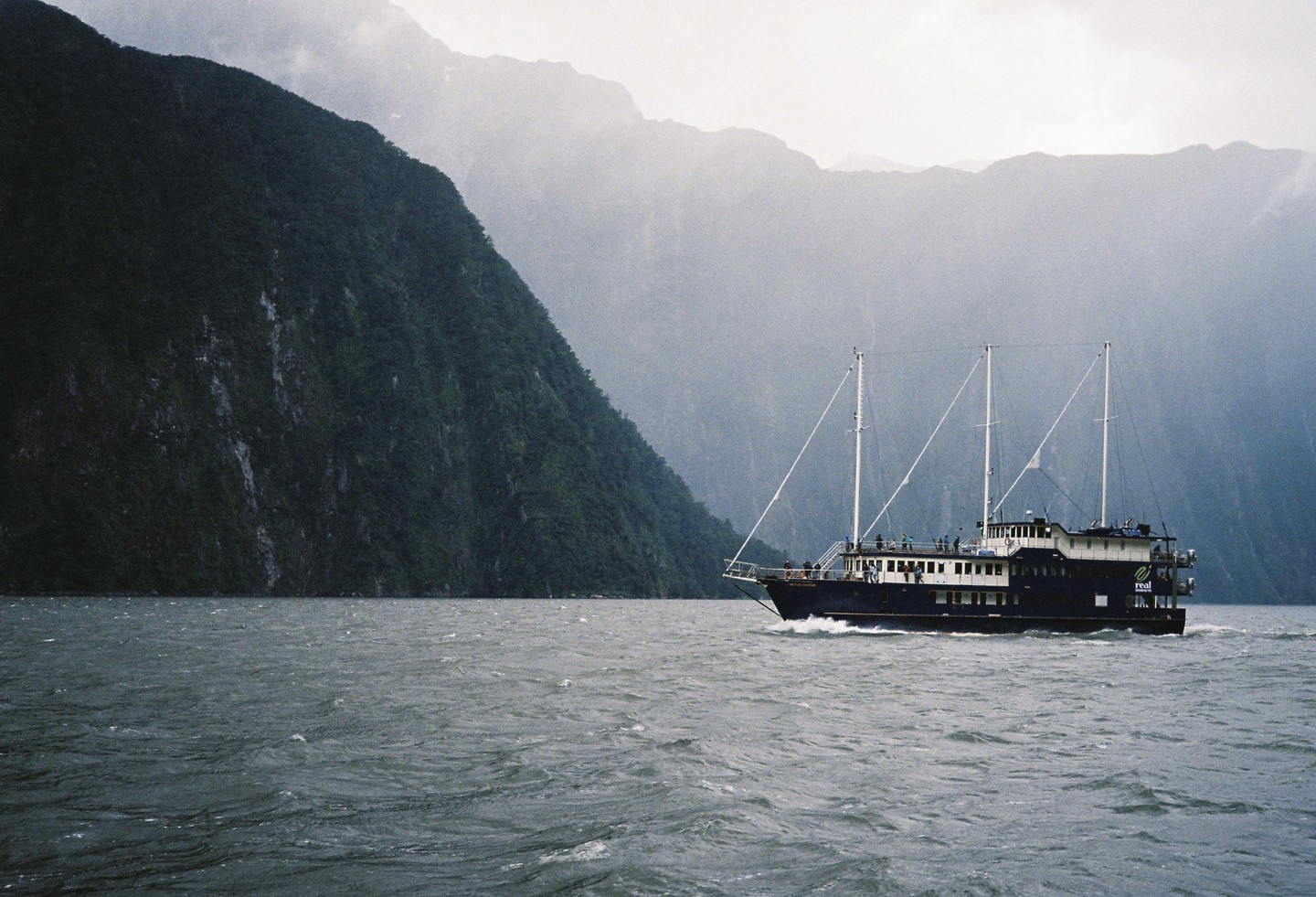 A boat with three masts travels on a body of water next to a large mountain.