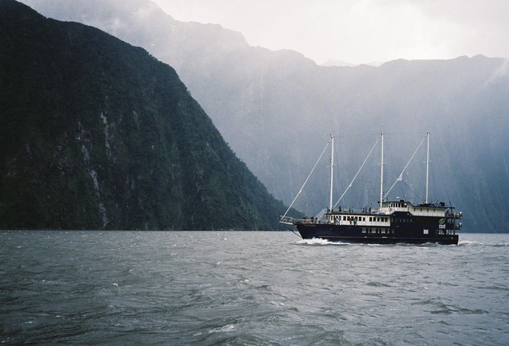 A boat with three masts travels on a body of water next to a large mountain.