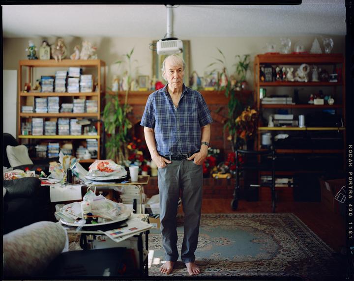 A man stands barefoot in a cluttered living room with shelves full of items and a patterned rug.