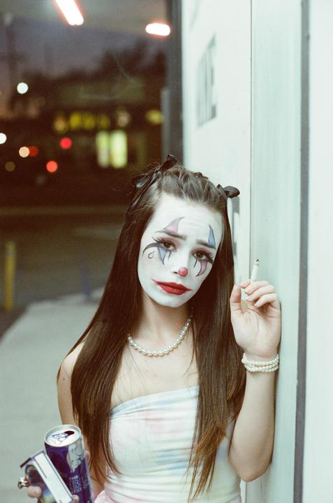 A woman with clown makeup, a pearl necklace, and a cigarette stands next to a wall at night.