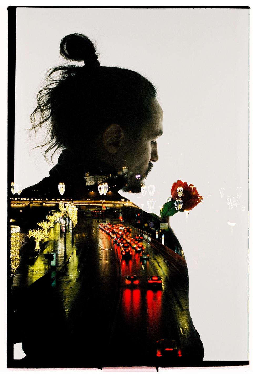 A man in profile with a rose is double exposed with a night street scene featuring car lights and illuminated trees.