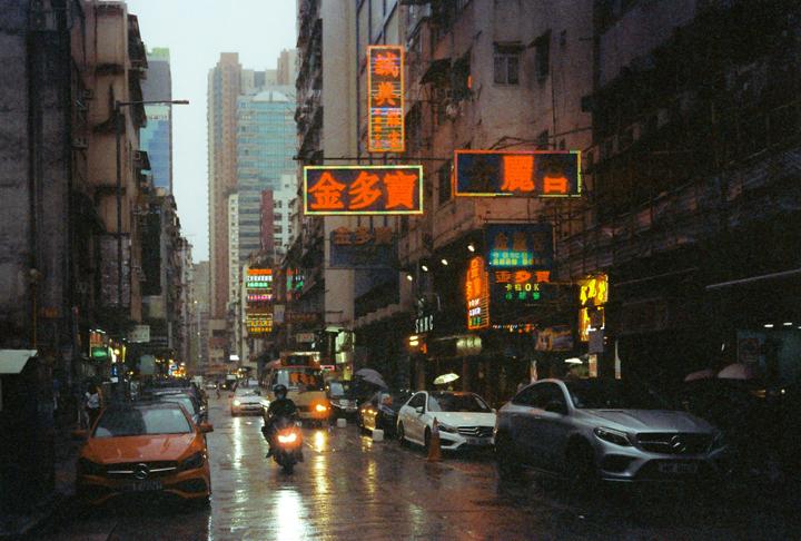 A street in a city is wet from rain, with cars and a motorcycle driving past buildings with neon signs.