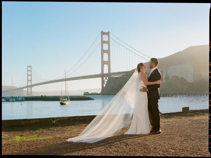 A couple embraces on a dirt path with the Golden Gate Bridge and a sailboat in the background.
