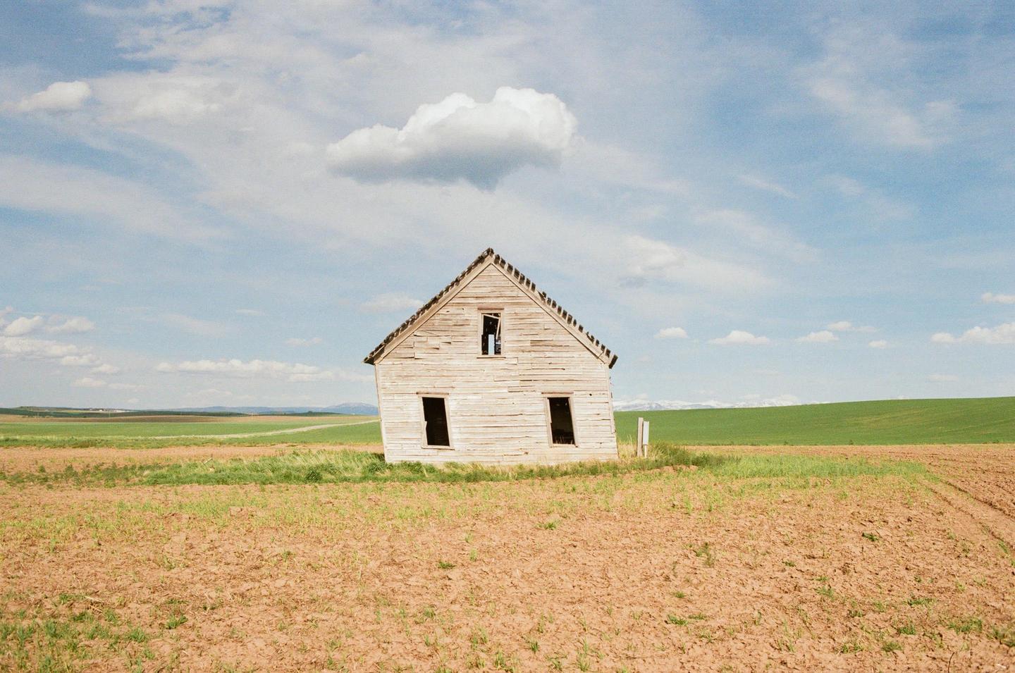 An abandoned house stands in a field under a partly cloudy sky.
