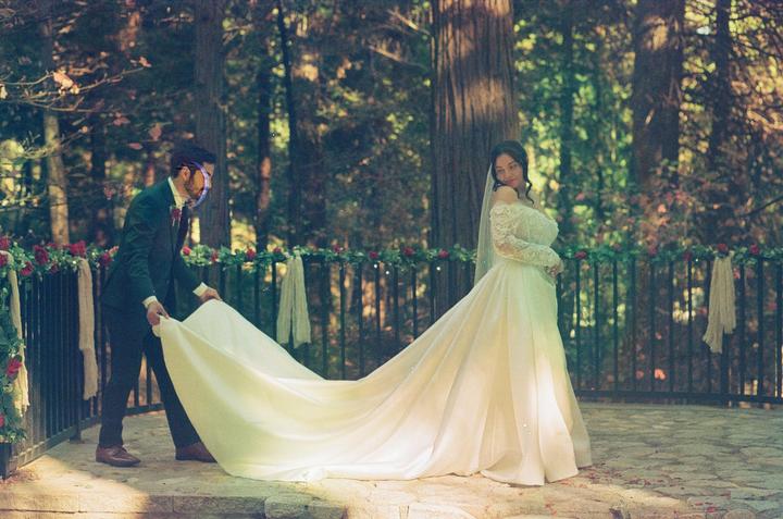 A man in a suit holds the train of a woman's wedding dress in a forest setting.