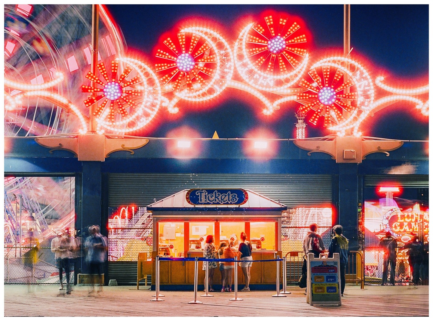 A ticket booth is brightly lit at night with neon lights and a ferris wheel in the background.