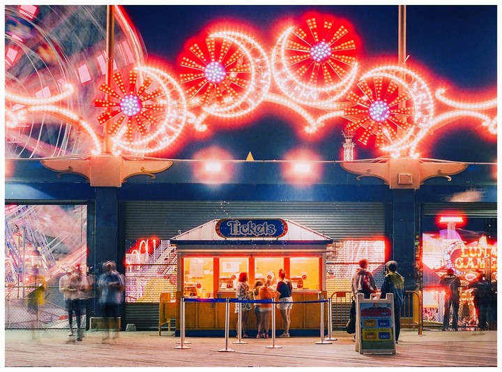 A ticket booth is brightly lit at night with neon lights and a ferris wheel in the background.