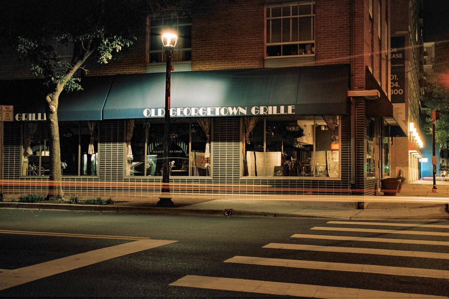 A street corner at night shows a restaurant with a blue awning and light trails from passing vehicles.