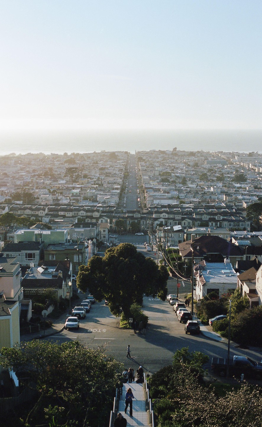 A long street extends through a dense residential area towards the ocean under a hazy sky.