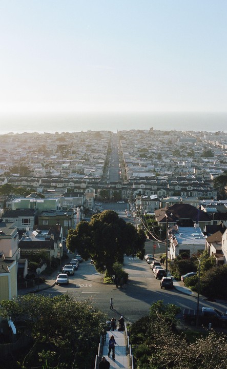 A long street extends through a dense residential area towards the ocean under a hazy sky.