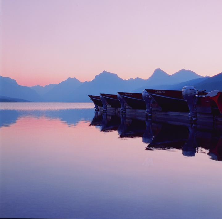 Several boats are docked on a calm lake with mountains in the background under a pink and blue sky.