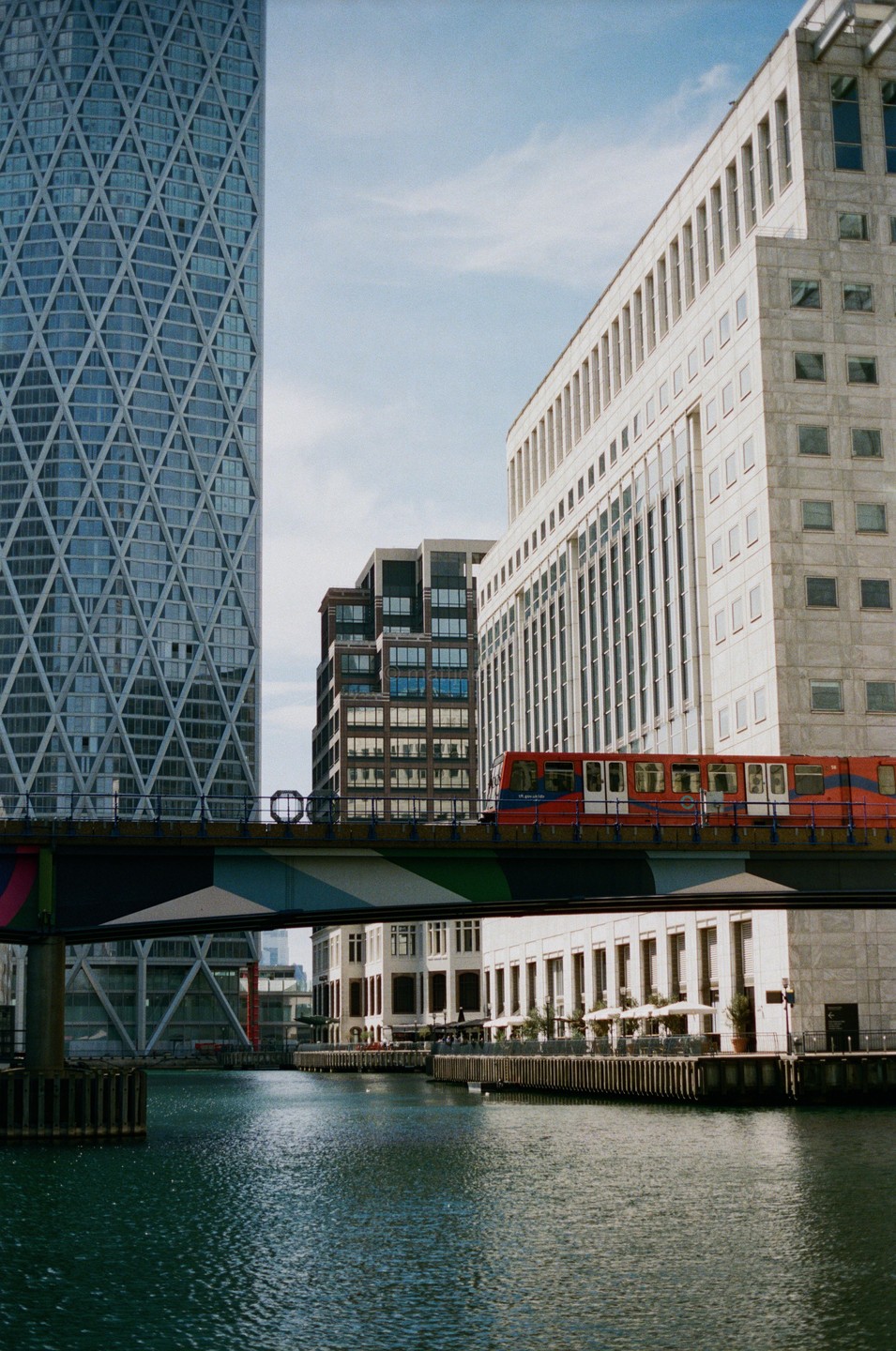 A red train crosses a bridge over a canal, surrounded by tall buildings in Canary Wharf.