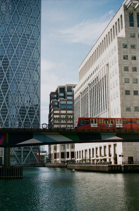 A red train crosses a bridge over a canal, surrounded by tall buildings in Canary Wharf.
