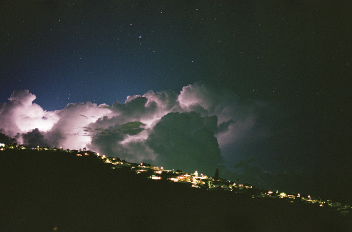 A lightning storm illuminates clouds above a hillside neighborhood at night.