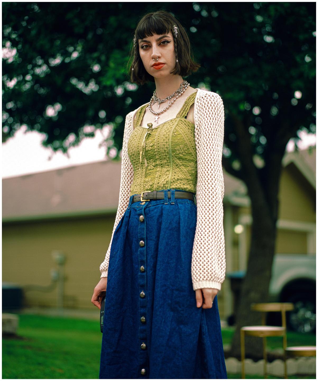 A woman with dark hair and bangs stands outdoors, wearing a green top, a blue skirt, and a white crocheted cardigan.