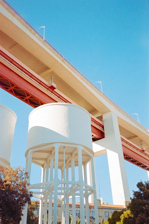 Two white water towers stand beneath a large bridge with red and white supports against a clear blue sky.