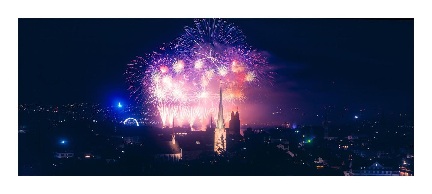Fireworks explode over a city skyline at night, illuminating a tall church steeple and other buildings.