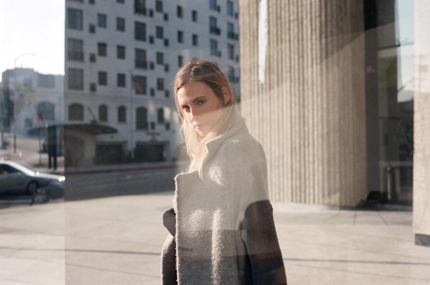 A woman in a color block coat stands in front of a building with reflections of the street and another building visible.