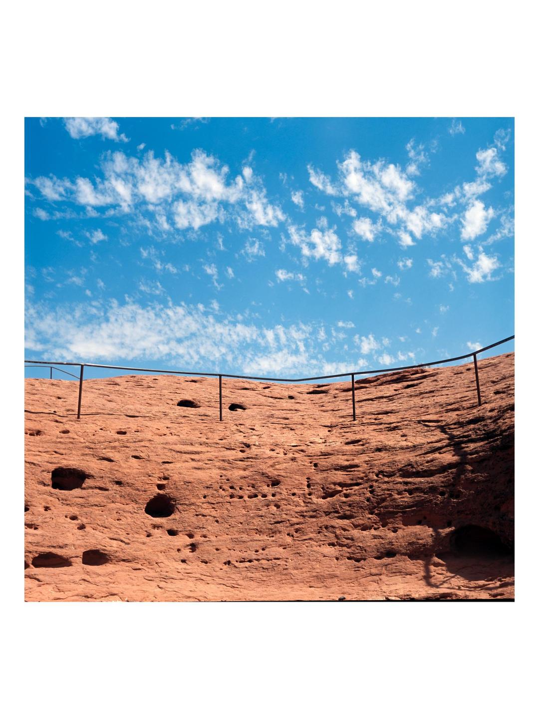 A metal railing curves along a textured red rock formation under a bright blue sky with scattered white clouds.