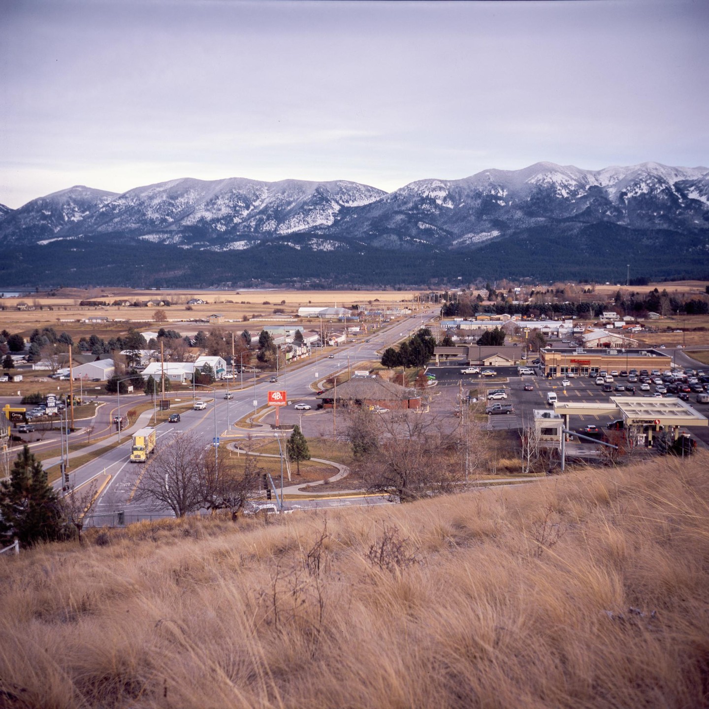 A small town with a highway running through it is nestled at the base of snow-capped mountains.
