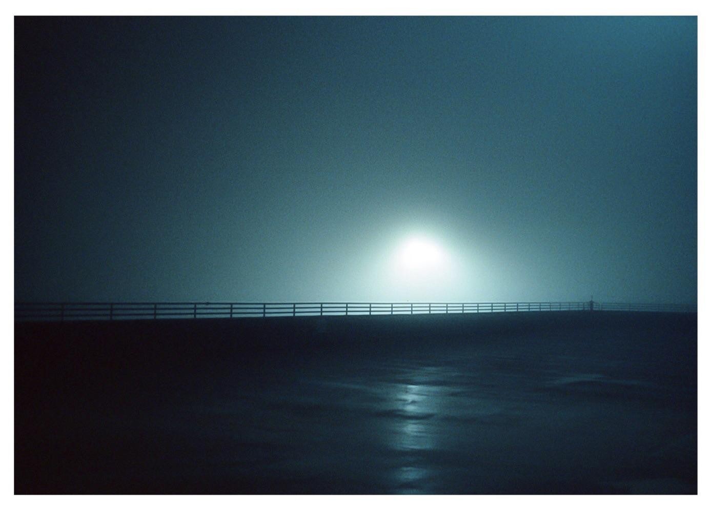 A bright light illuminates a fence and a wet surface under a hazy sky.