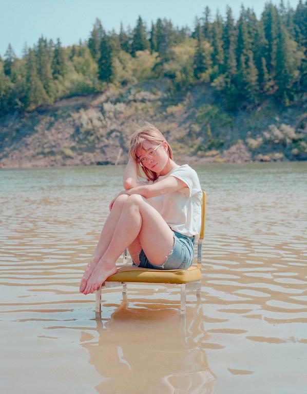 A woman sits on a chair in shallow water with a forest in the background.