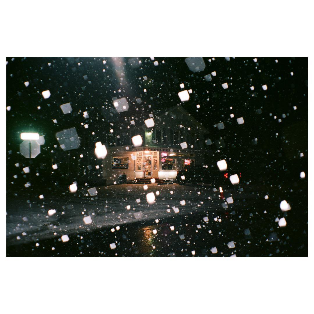 A building with illuminated windows is seen through falling snow at night.