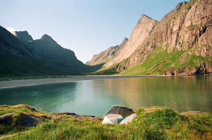 A tent is pitched on a grassy bank overlooking a calm body of water surrounded by mountains.