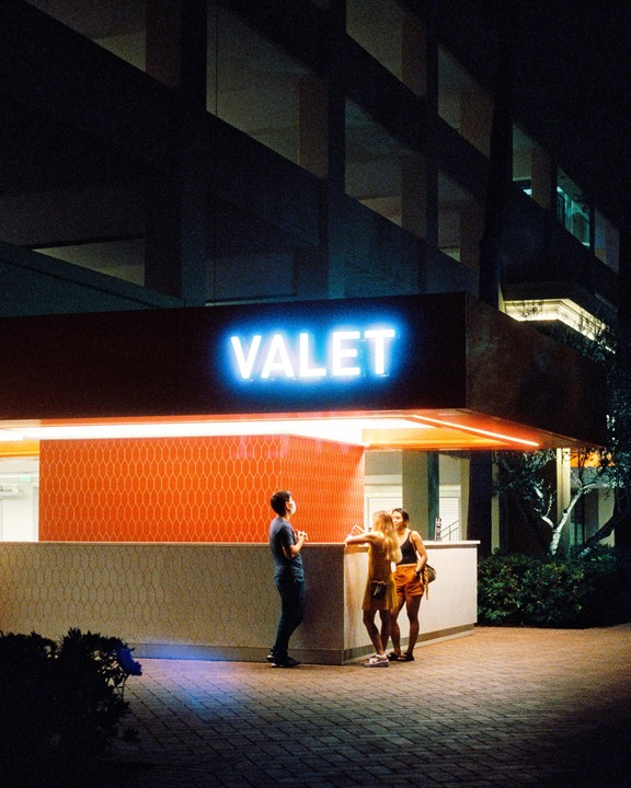 Three people stand near a valet stand with a bright blue neon sign at night.
