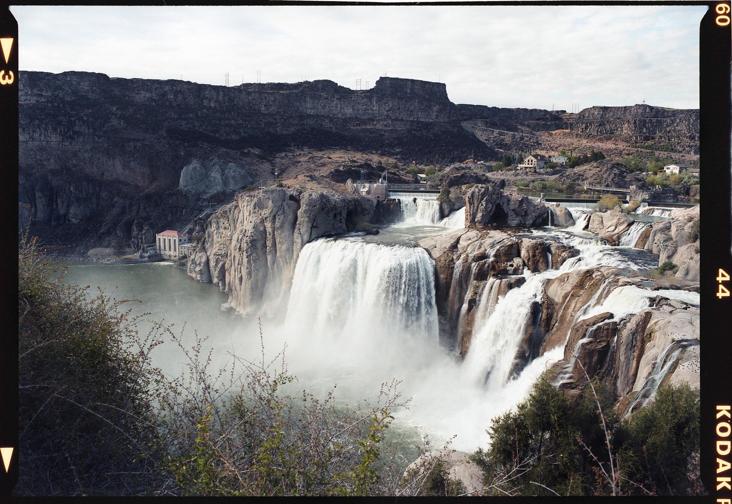 A wide waterfall cascades over a rocky cliff into a river, with a building visible on the far bank and a rugged landscape in the background.
