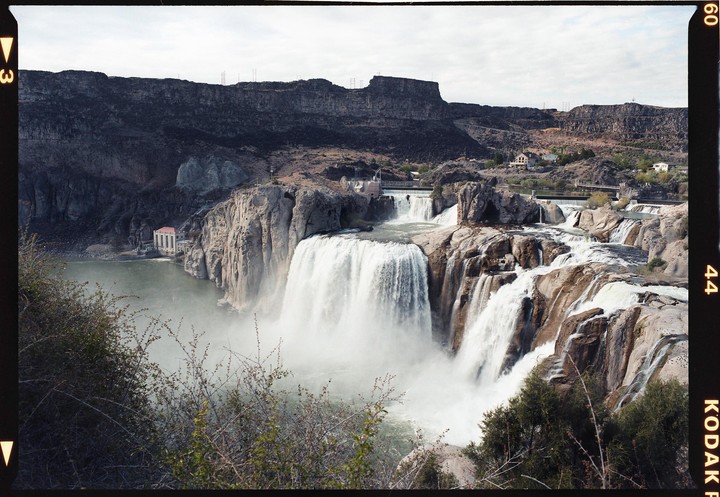 A wide waterfall cascades over a rocky cliff into a river, with a building visible on the far bank and a rugged landscape in the background.