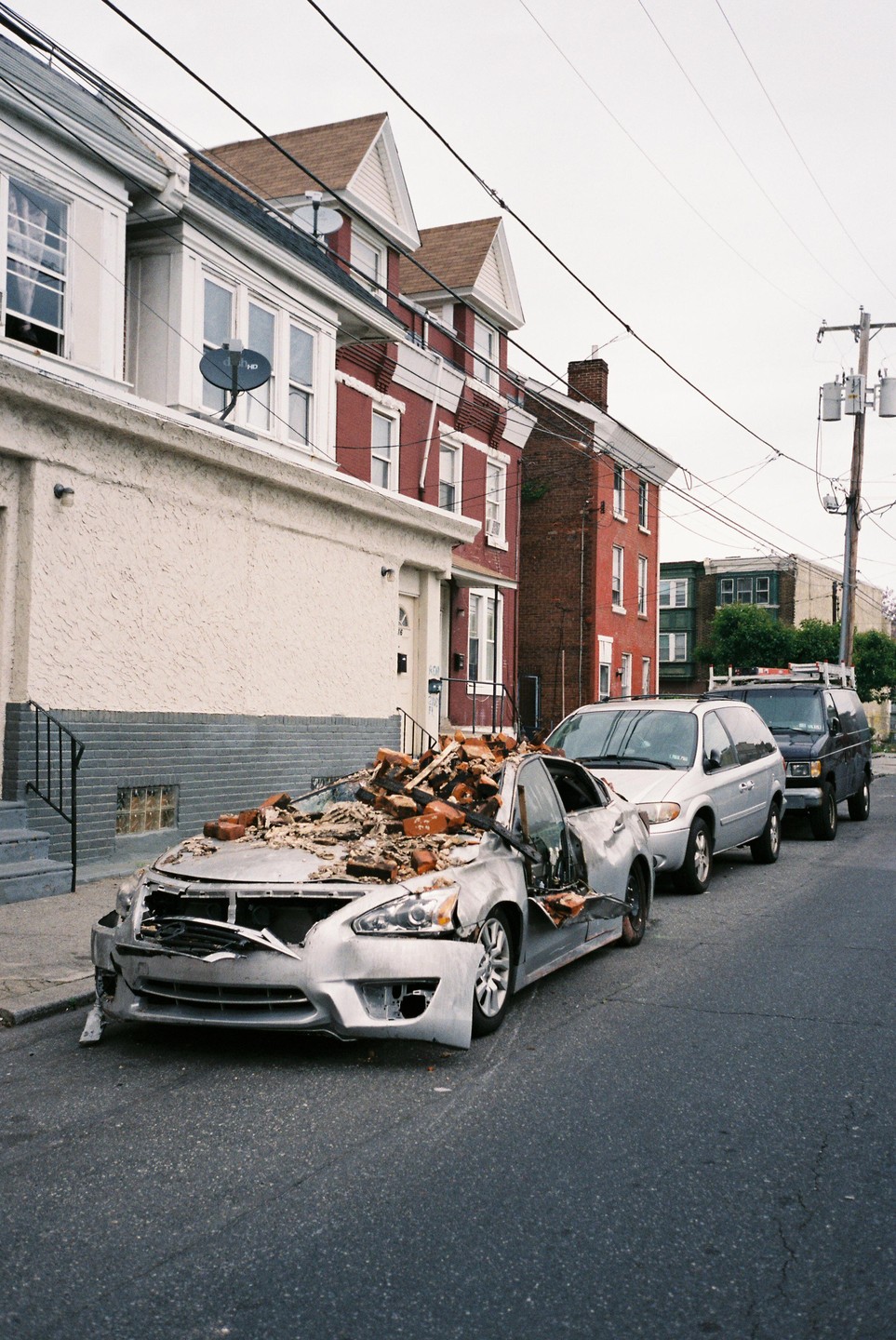 A silver car is parked on a street with its roof caved in and covered with bricks and debris.