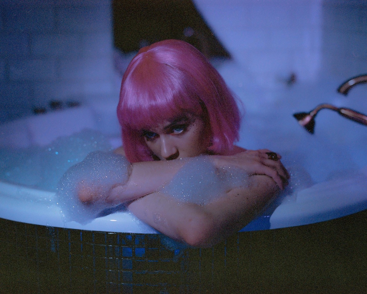 A woman with pink hair and blue eyes looks out from a bubble bath.