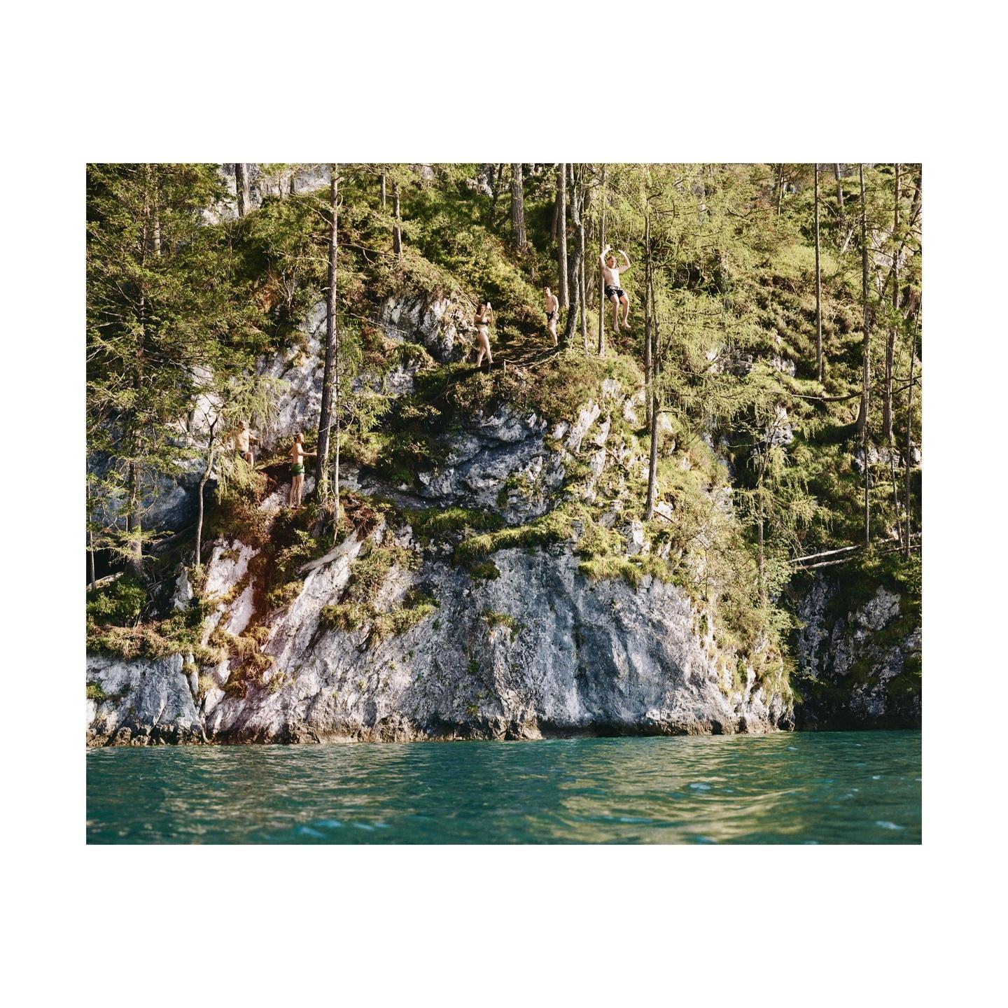 A group of people are on a rocky, tree-covered cliff overlooking a body of water, with one man swinging from a rope.